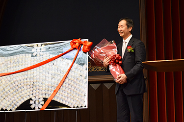 Professor Kajita smiles in front of a postcard-decorated board as he holds a bouquet, both presented to him by the University. Written on the board's small postcards featuring photos of the Super-Kamiokande's photomultiplier tubes are messages from University members