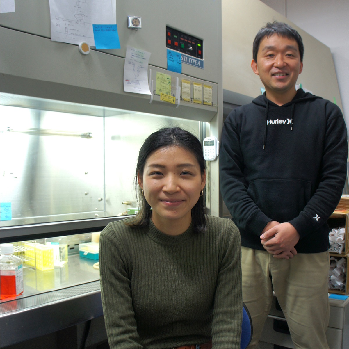 Two scientists in a laboratory, one standing and one sitting infront of a cell culture hood. 