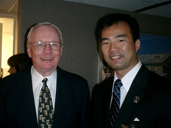Neil Armstrong (left) stands next to Soichi Noguchi (right). Both men are wearing suits and smiling.