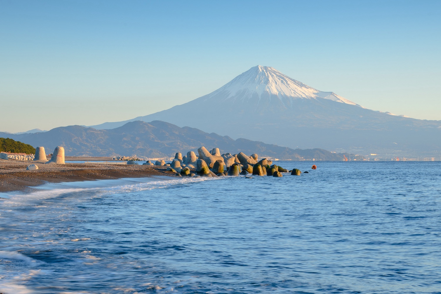 A photo from the coast of mount fuji on a sunny day, with tertrapods piled on the beach in front.