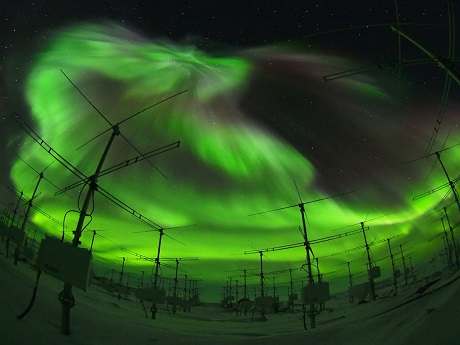 Bright green aurora lights up the night sky behind the dark outline of radar antenna