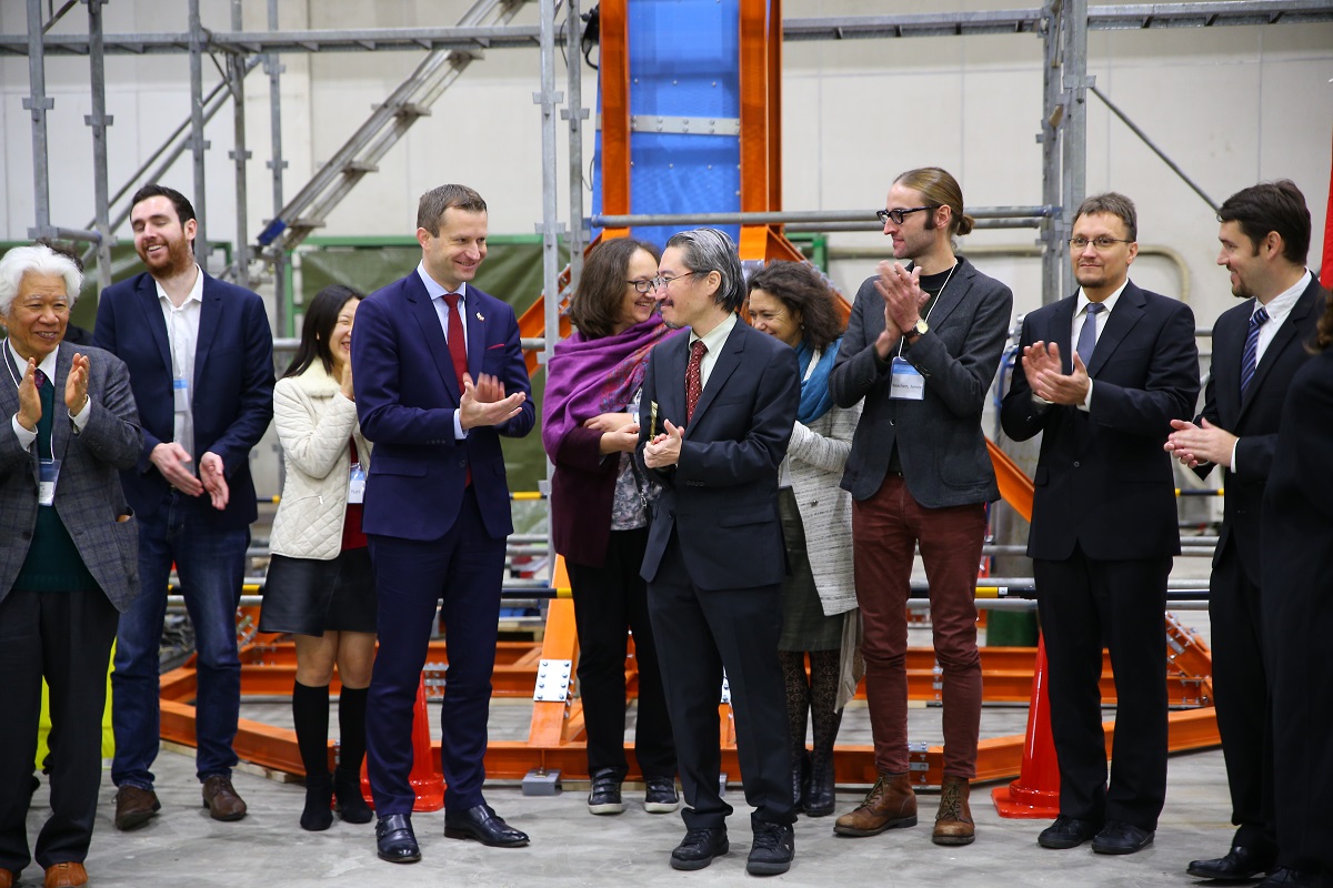 A group of people dressed smartly stand in front of a blue machine clapping their hands