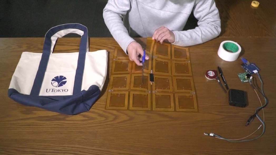 On a desk there is a bag then an orange sheet with electronic components on it being cut with a pair of scissors