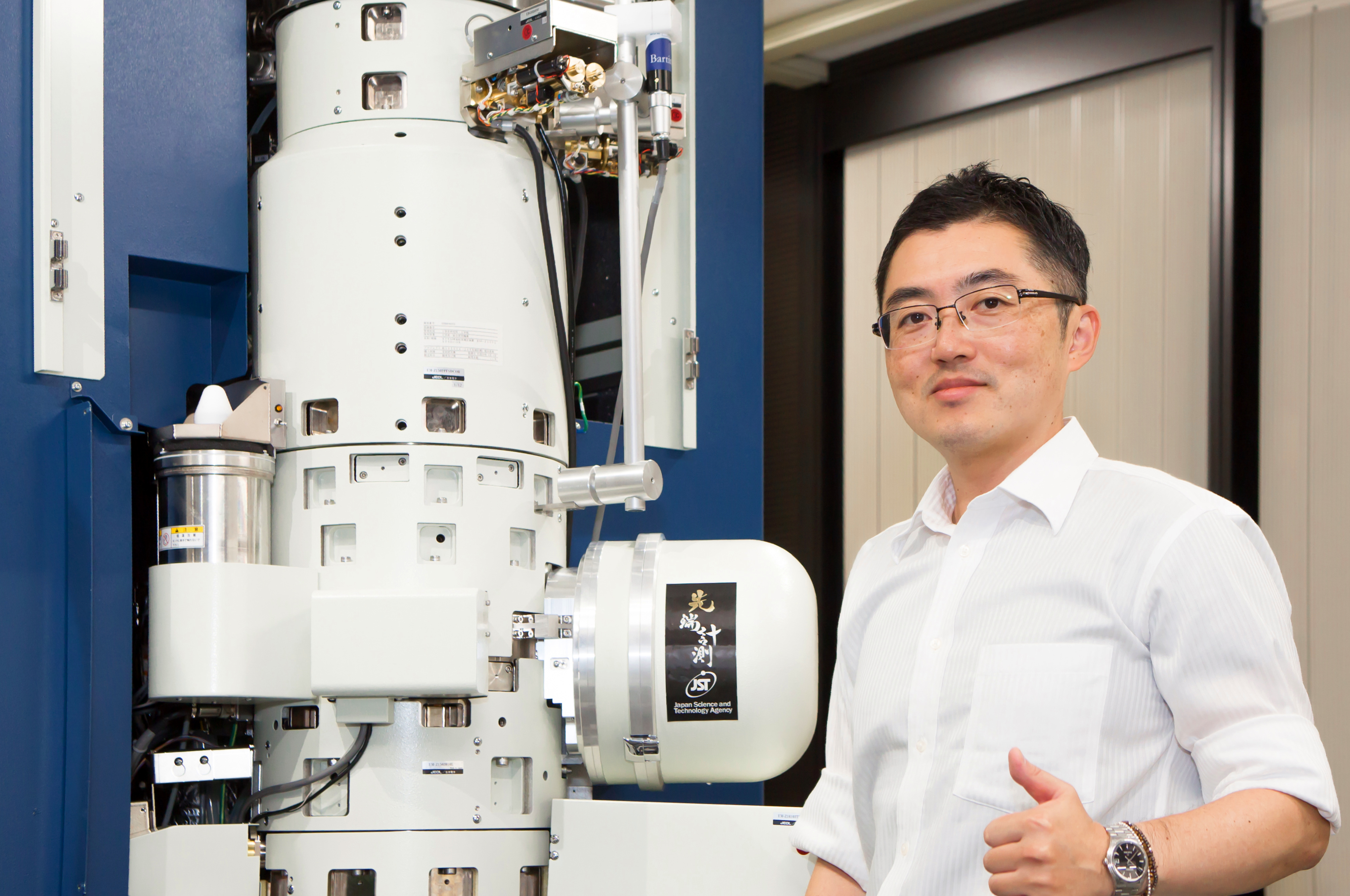 On the left is an electron microscope, a large machine made of white cylinders and covered in electronic components. On the right is a man, Professor Shibata.