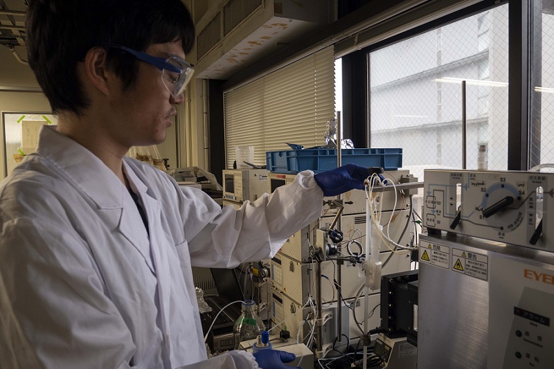 A male researcher wearing a white coat and blue gloves operates a piece of laboratory equipment in front of a window.