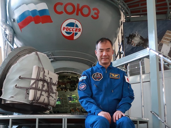 Soichi Noguchi wears a blue jumpsuit and sits in front of the Soyuz Simulator, which is grey and round.