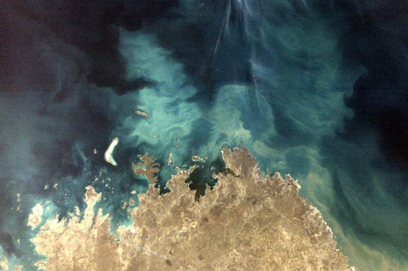A brown tip of land at the bottom of the image is surrounded by light and dark blue swirls of ocean in the rest of the image.