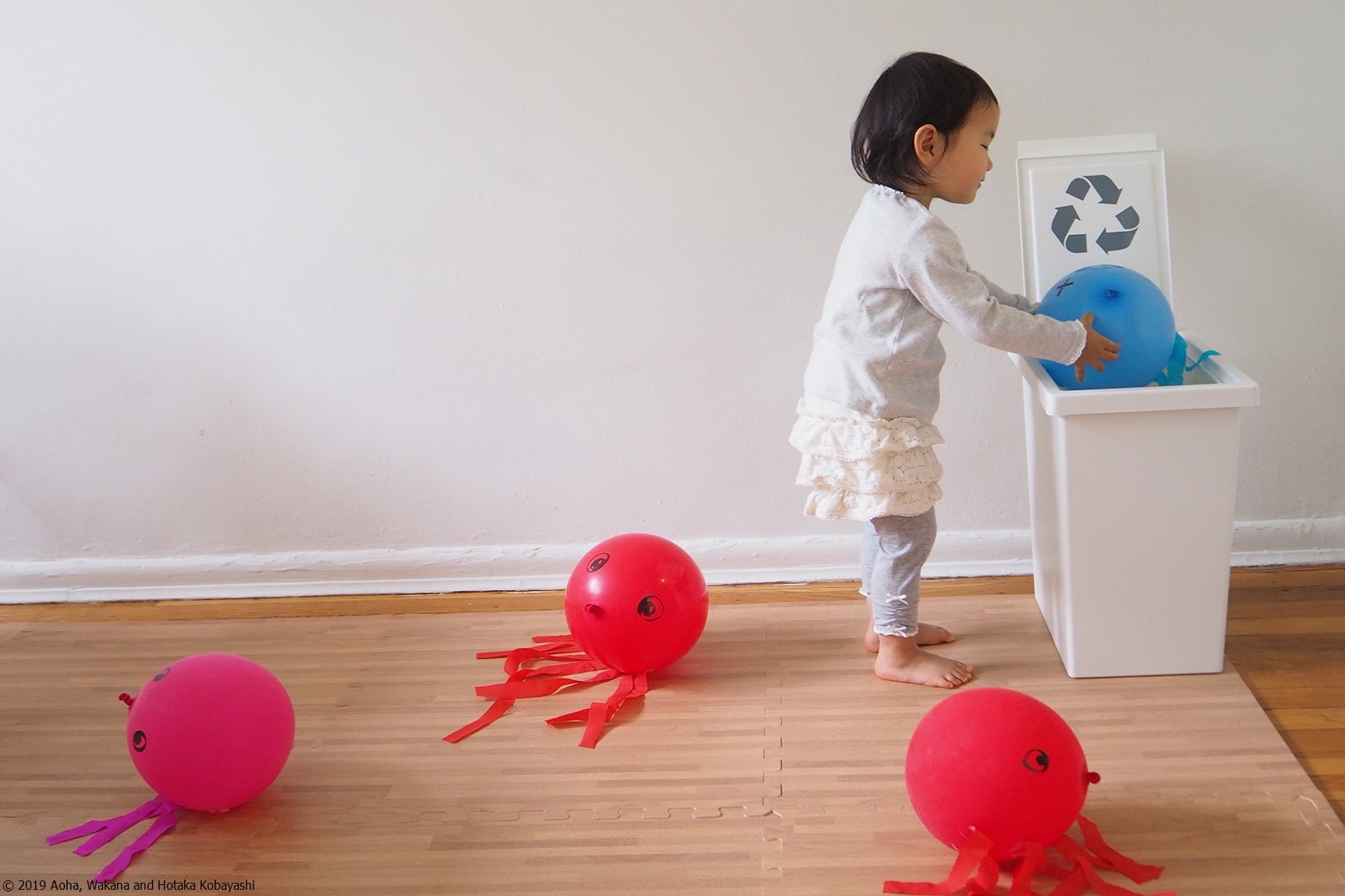 A little girl places a blue, octopus-shaped balloon into a recycling bin while red balloons rest on the floor.