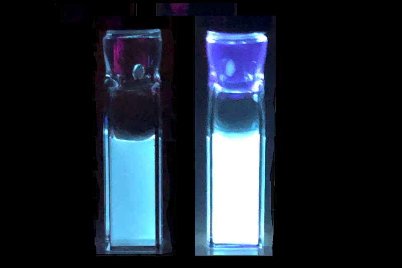 Two rectangular containers filled with liquid glowing blue under UV light. The container on the right side is approximately 3.9 times brighter than the one on the left.