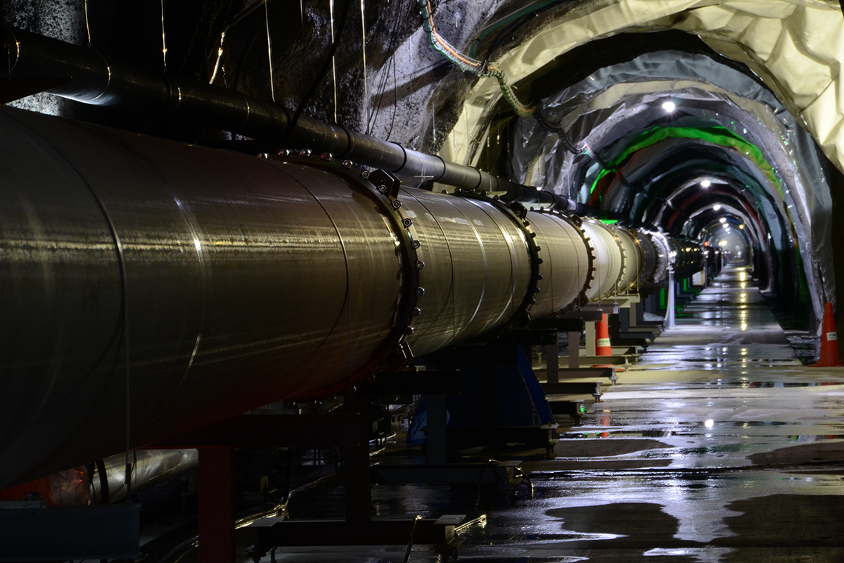 A tube inside a dimly lit underground tunnel spans off into the distance. Overhead electric lights reflect in small puddles on the ground.