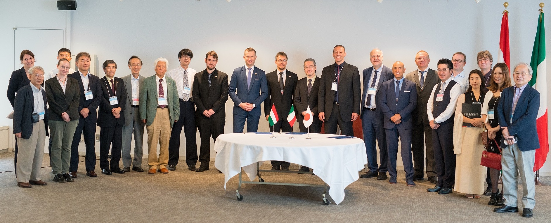 A group of people standing behind a table on a grey floor. On the right the Italian and Japanese flags hang on poles behind the crowd.