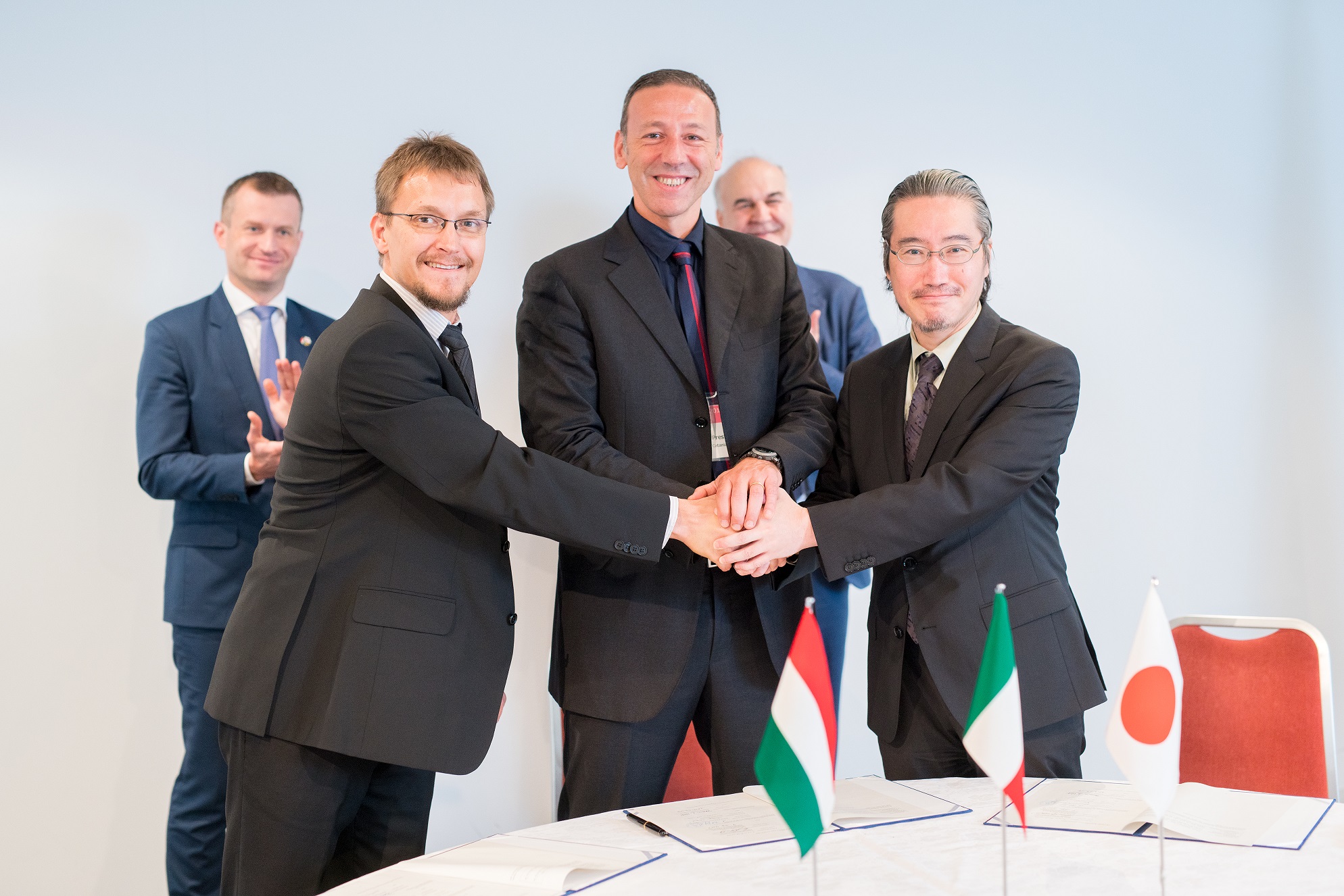 Three men in the foreground shake hands over a white table. There are documents on the table.