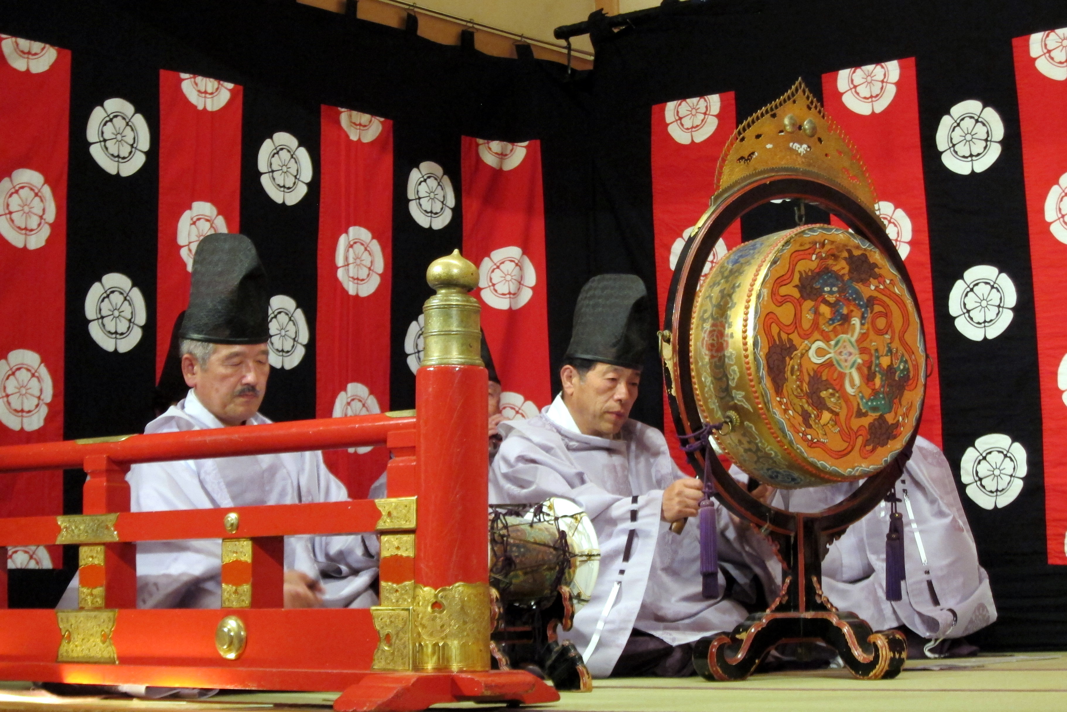 A red and black background. Three men in light robes play ancient instruments.