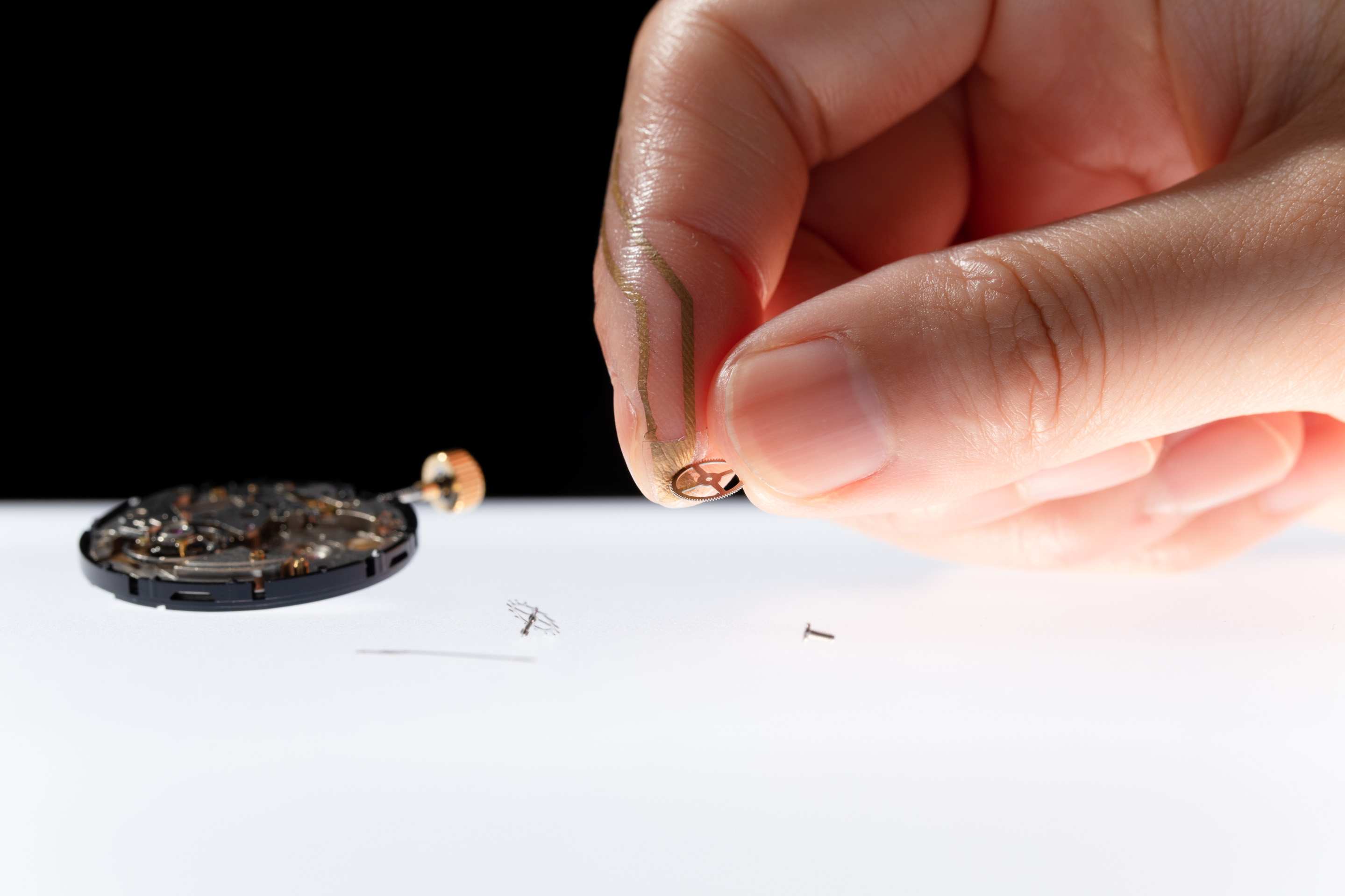 A black background. A close up of two fingers holding a cog. A white surface is below.