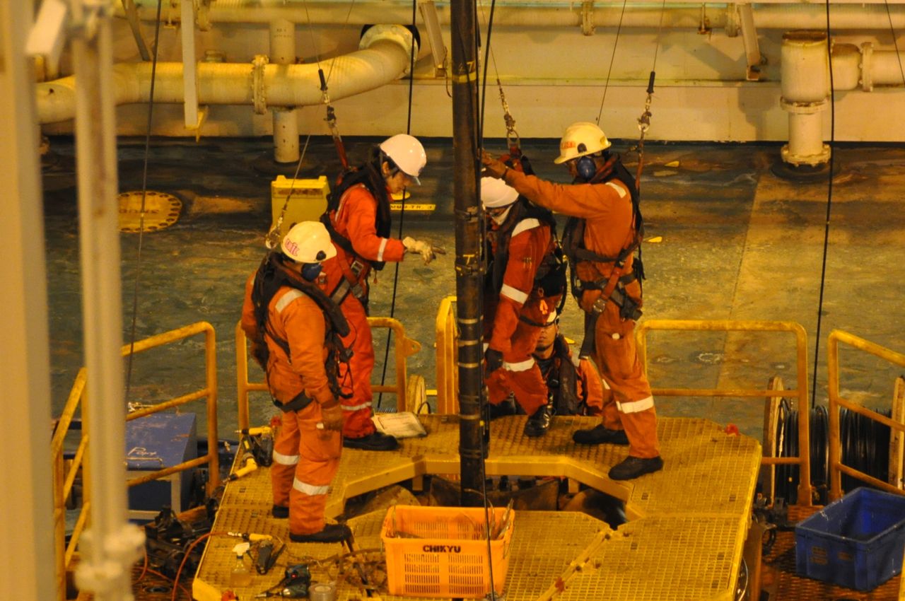 A group of people in overalls surrounding a vertically orientated metal shaft