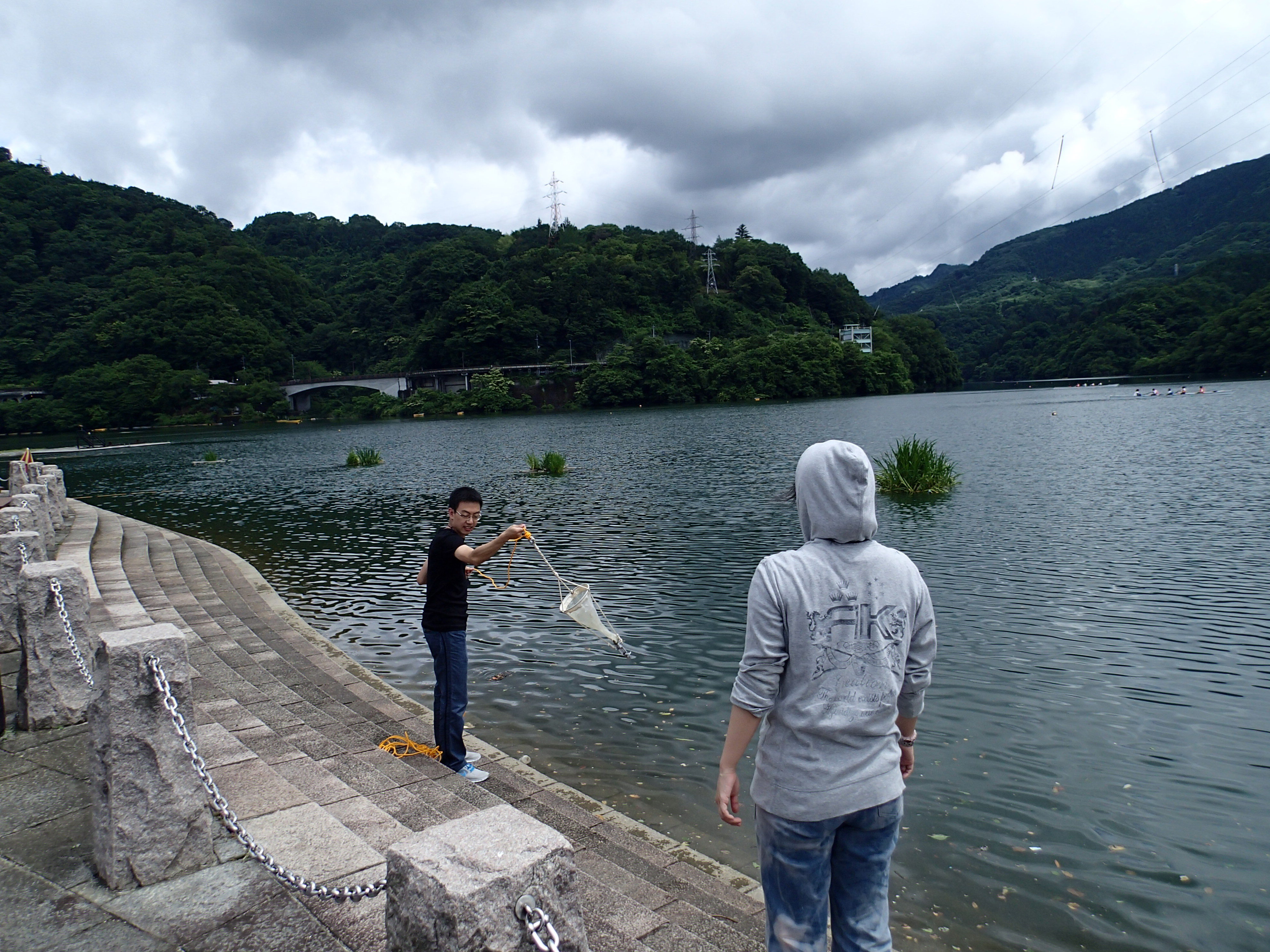 Two young men in casual clothes standing on cement steps at the edge of a body of water. One swings a algae collection net.