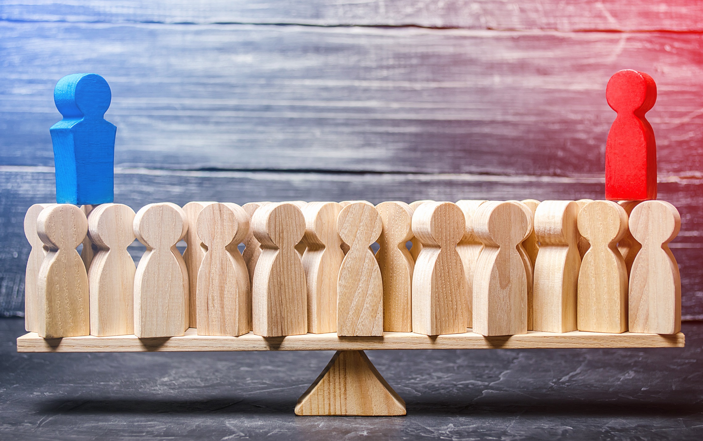 A row of wooden figures on a see-saw, a blue one on the left and a red one on the right stand atop the rest.