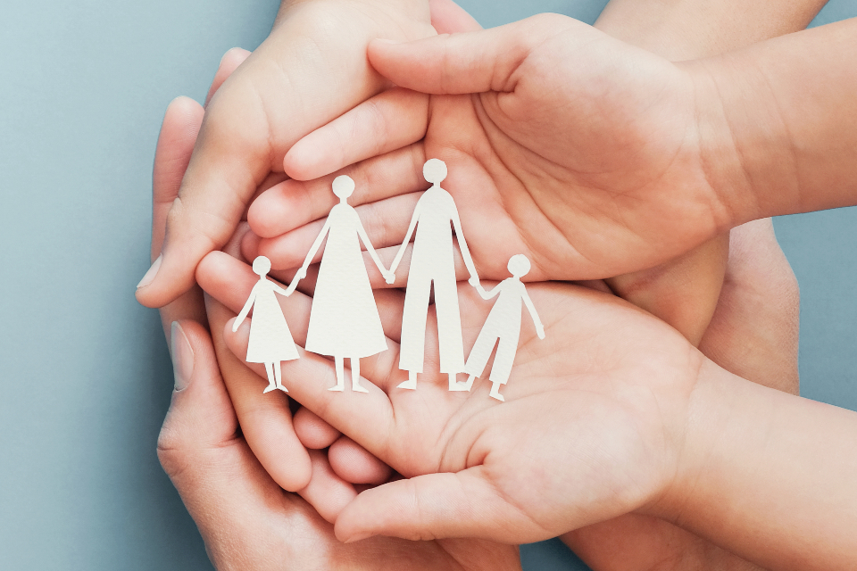 Photo of three people’s hands stacked on top of each other, palms up, holding a white paper cutout silhouette artwork of a family of four (son, father, mother, daughter) standing in a line and holding hands.