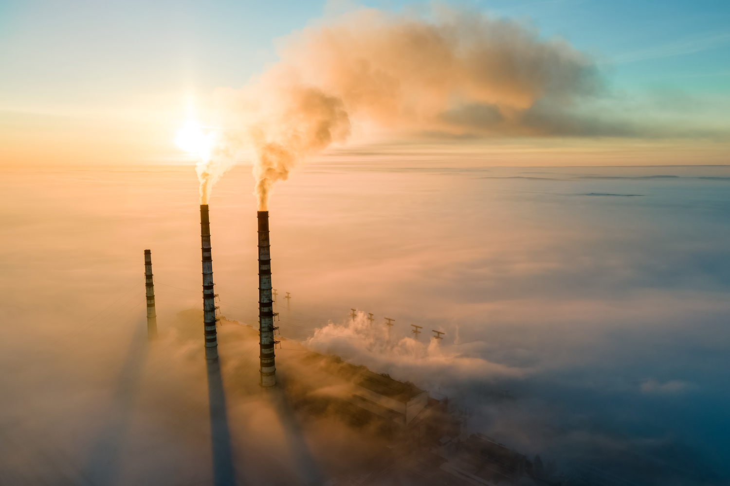 Coal power plant chimneys tower above low clouds, pumping steam into the blue sky at sunrise.