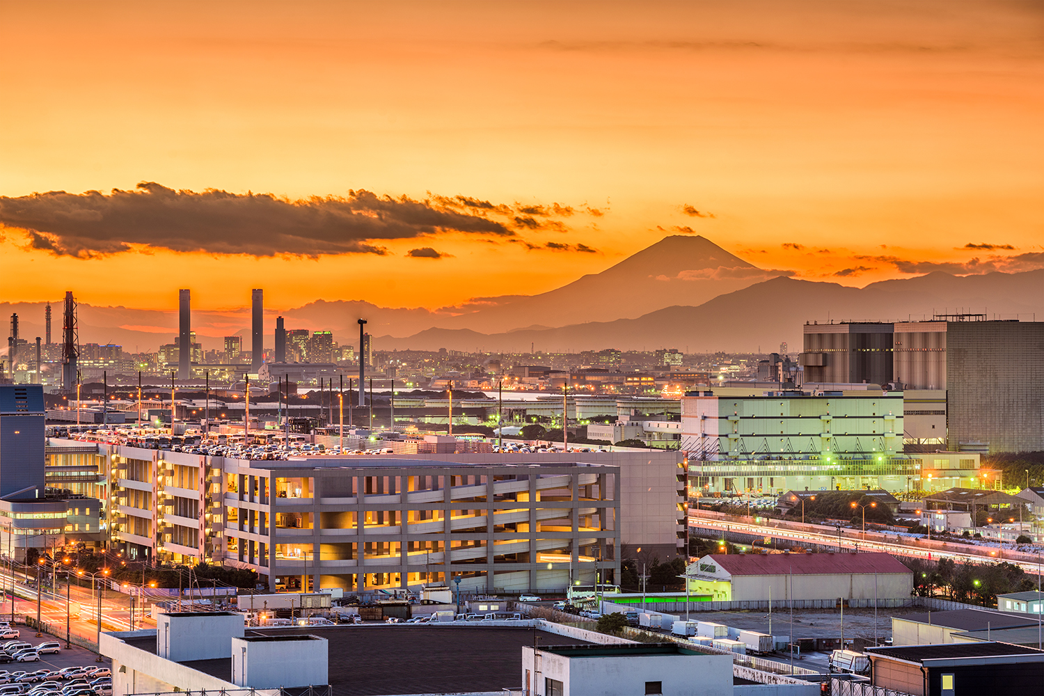 Japan in an orange sunset, mount fuji towers in the distance behind a factory district.