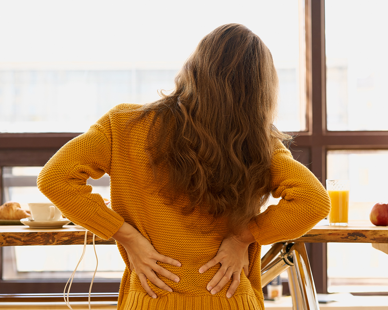 Back view of young woman with long red hair holding lower back in discomfort.