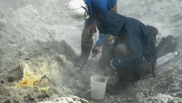 Short video of several people wearing protective gear and collecting fumarole gases, first close up and then from a distance, with steam and gas spewing from the fumaroles around them.