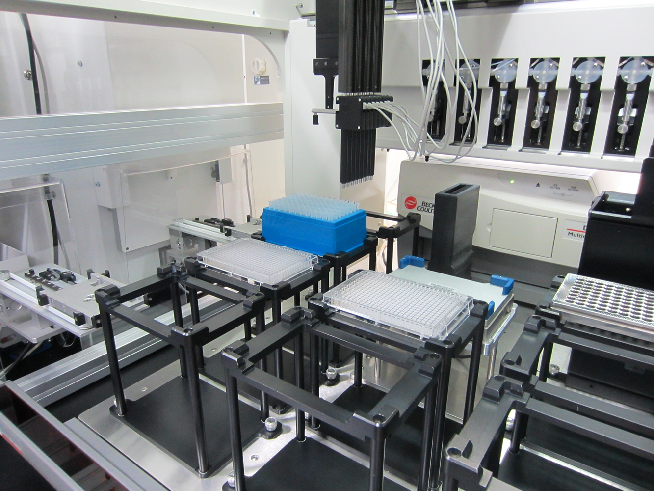 In a lab, a robot arm hangs from the ceiling while multiple trays for the experiment have been positioned below.