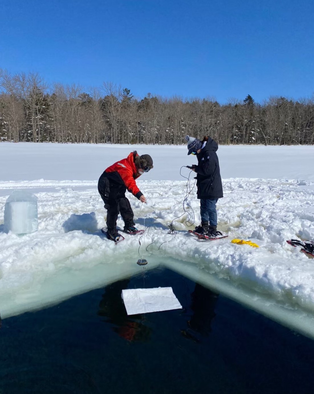 Two researchers drop a measuring device into a large square hole in the snow-covered ice, on a cold and bright sunny day, on Lake Akan.