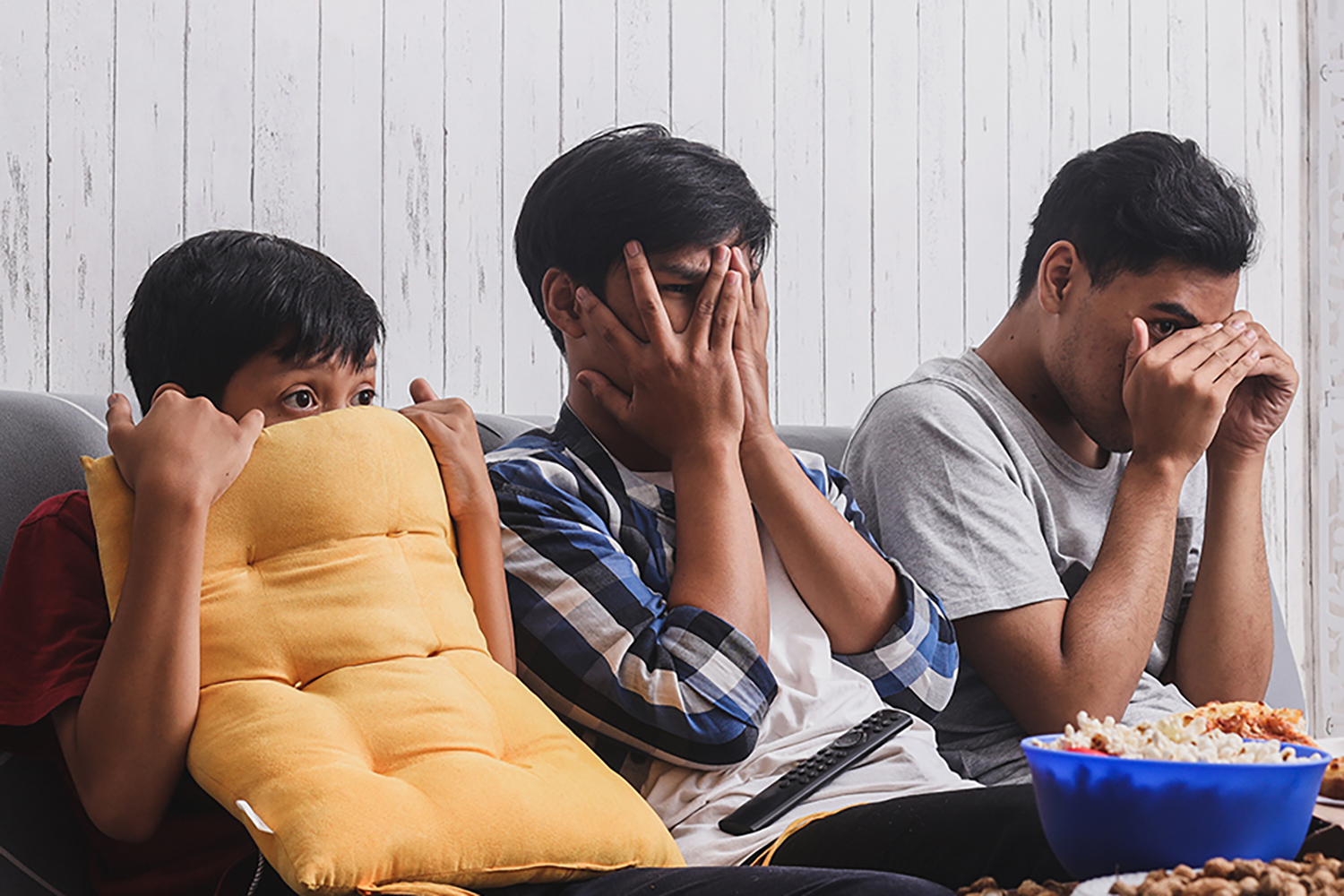 A photography of three boys and young men hiding their eyes while watching a scary movie.