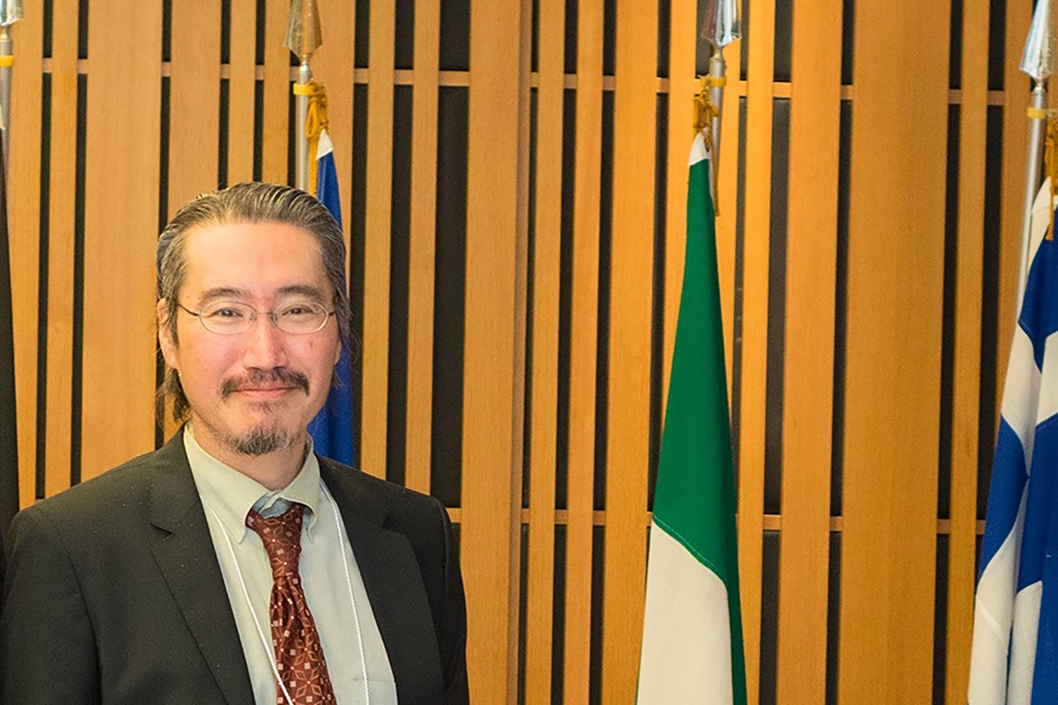 A portrait photo of Professor Hiroyuki Tanaka, who is smiling and wearing a suit while standing in front of some flags in a wood pannelled room. 