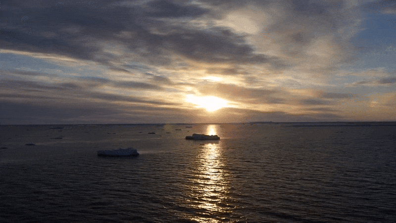 Video flying over antarctic ocean with clouds and setting sun reflected on the sea.