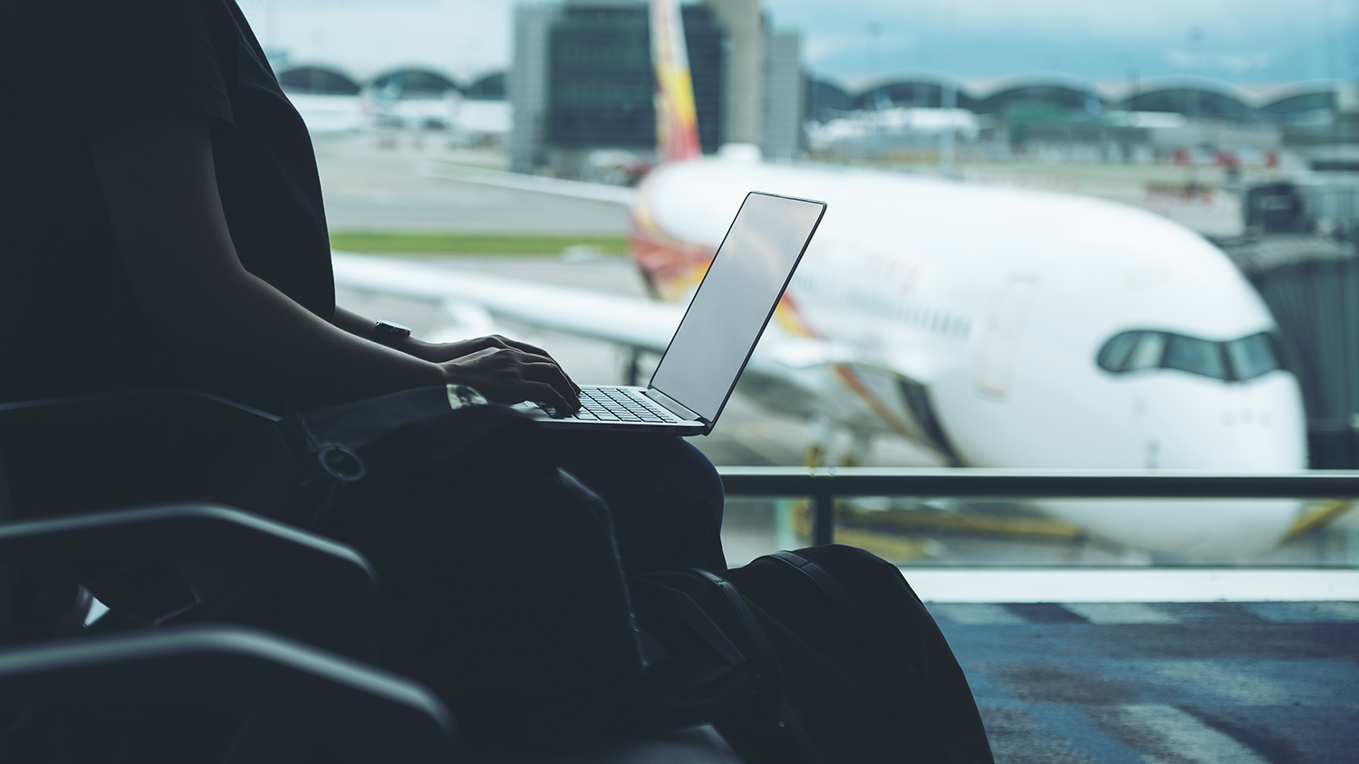 Photo of back illuminated woman sitting at an airport waiting area typing on a laptop, with a plane docked in the background.