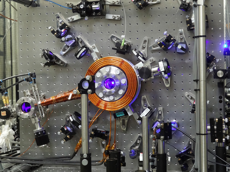 An optical lattice clock. A porthole with a diameter of 30 mm offers a glimpse into the ultra-high vacuum chamber, where approximately one million laser-cooled ultracold strontium atoms emit bluish-white fluorescence. Researchers trap these atoms in an optical lattice constructed by the Magic Wavelength, measure their resonant frequency, and use it as a clock pendulum. © Hidetoshi Katori