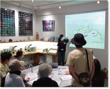 「サケの旅」と書かれたオホーツク周辺の地図が表示されたスライドの説明をする職員と見学者