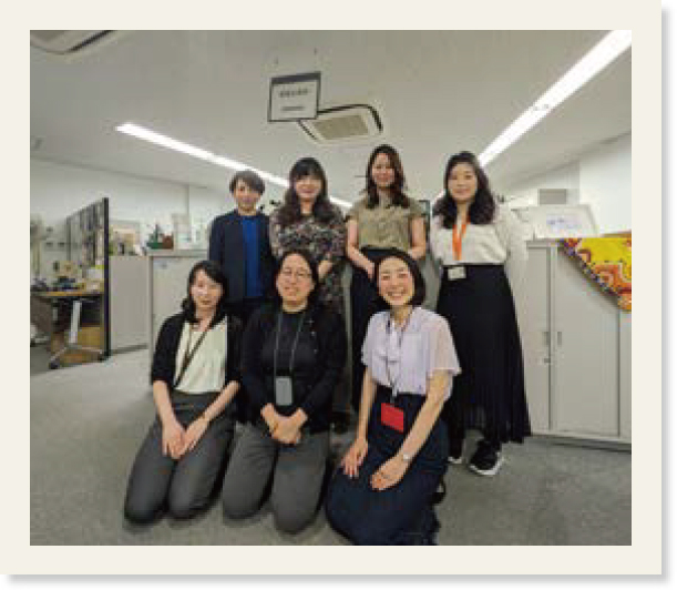 「経営企画部 国際戦略課」と書かれた吊り下げ看板の手前で写るチームの集合写真