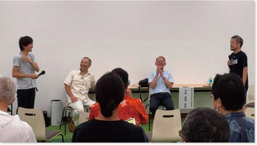 白い壁際の長机の手前に教授4人が並んで話をする様子と、向かい合って座る参加者