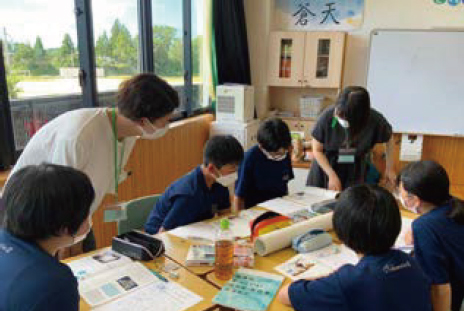 教室で机をコの字型に並べて座る子供5名と両脇に立つ先生2名。1人の子の冊子を見ている様子