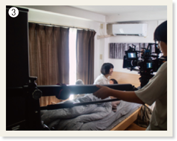 ❸部屋のベッドで寝ている男性と起き上がっている女性。その周りにカメラなどの撮影機材がある