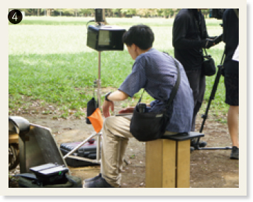 ❹公園のような場所で木の椅子に座ってモニターを見ている宮坂さん