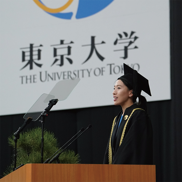 東京大学のロゴのパネルを背景に壇上から観客席の方を見て祝辞を述べる米田あゆさん