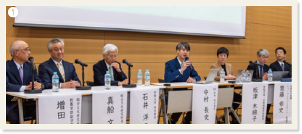 ❶スクリーンの下で名前の紙が貼り付けられたテーブルに座っているパネル1の7人の登壇者
