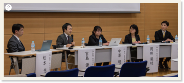 ❷スクリーンの下で名前の紙が貼り付けられたテーブルに座っているパネル2の5人の登壇者