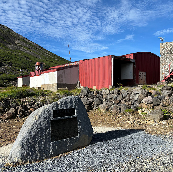 山のふもとにある赤い建物とその手前の「朝日の小屋」と書かれた碑