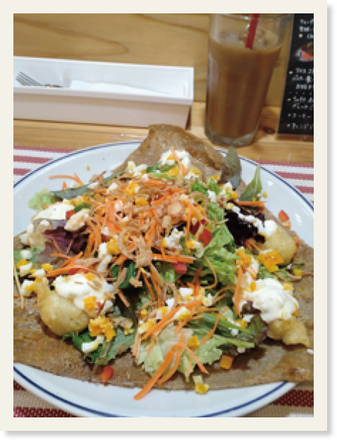 白いお皿に乗っている野菜とフリッターのガレット（手前）とグラスに入った飲み物（奥）