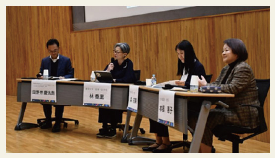 壇上のスクリーン前の長机に座る座談会の登壇者。うち1人がマイクを持って話している様子
