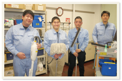 水色の作業服を着た4人がそれぞれ掃除道具を持って集合写真に映る様子