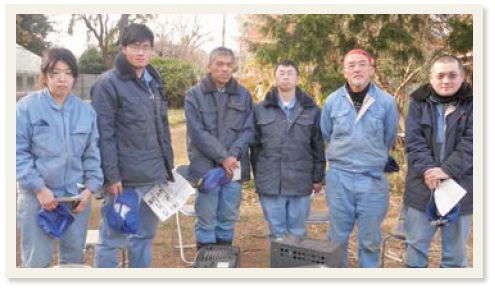 青色の作業服を着た6人がそれぞれ作業道具や帽子などを持って集合写真に映る様子