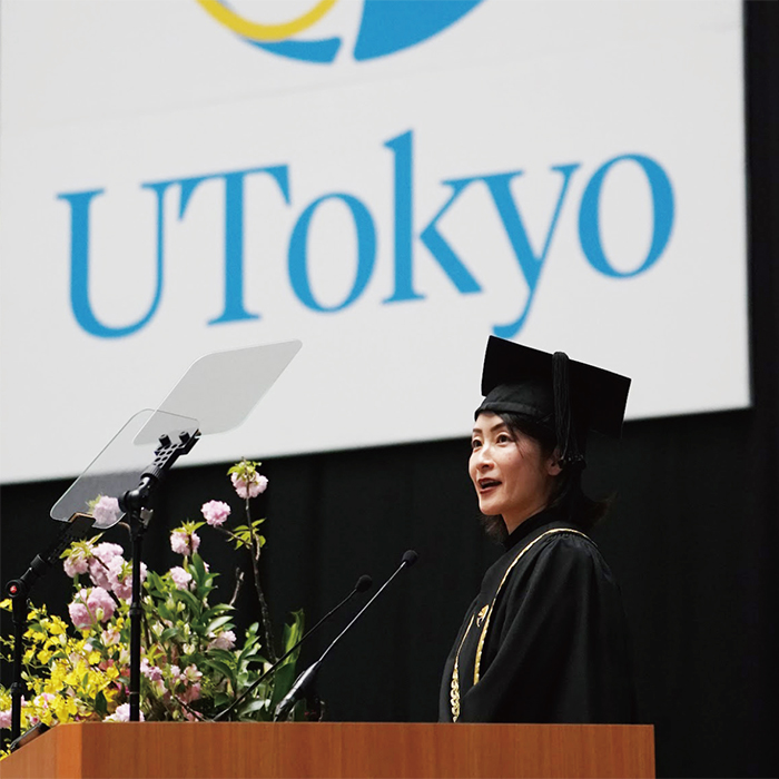 東京大学のロゴのパネルを背景に壇上から観客席の方を見て祝辞を述べる土井香苗さん