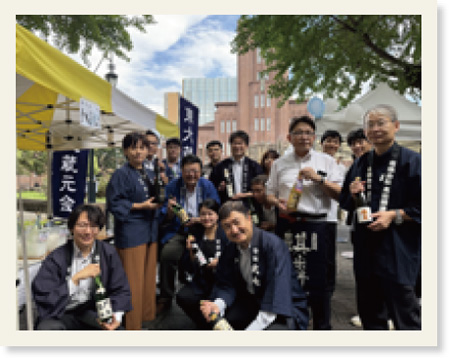 酒瓶を持つ人々が笑顔でポーズをとる写真、銀杏並木の利き酒会の様子