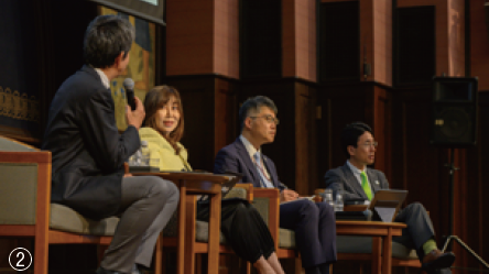 ②4人が座って登壇し1人がマイクを握っている様子